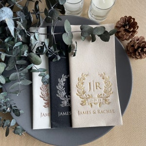 May include: Three white napkins with gold foil designs. The napkins feature a wreath with the initials "J R" and the date "25 09 23". The text "JAMES & RACHEL" is printed below the wreath. The napkins are on a black plate with eucalyptus leaves.