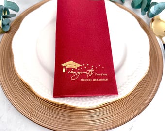 May include: A rectangular deep red paper napkin featuring gold foil printing. The design includes a graduation cap icon, a trail of small stars, the word "congrats" in script, "Class of 2025", and the name "KIDOUSE MEKONNEN" in capital letters.