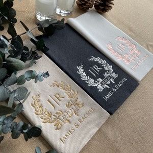 May include: Three linen napkins with gold, silver, and pink embroidery. Each napkin features a wreath with the initials "JIR" and the date "25.09.23". The text "JAMES & RACHEL" is printed below the wreath.