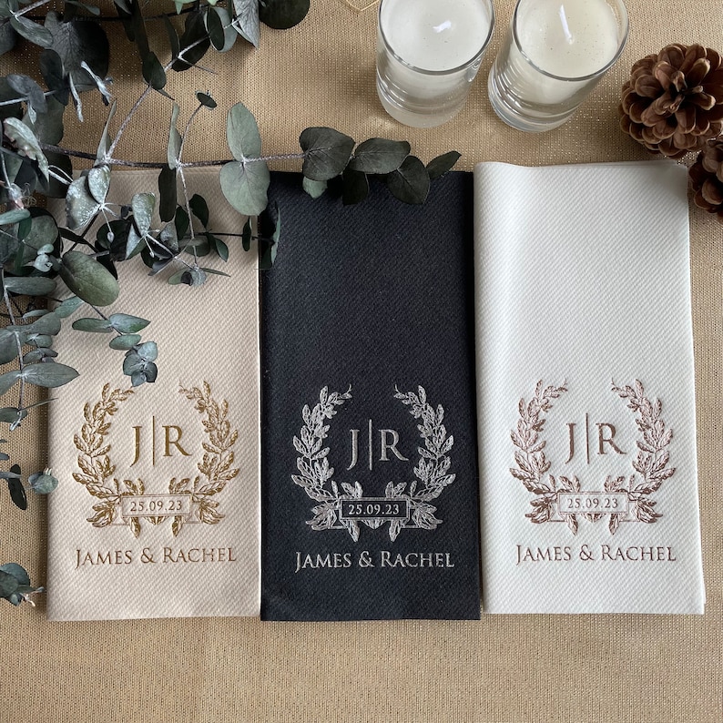 May include: Three white napkins with a gold, black, and silver design. The design features a wreath with the initials "JR" and the date "25.09.23" on a banner. The text "JAMES & RACHEL" is printed below the wreath.