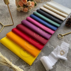 May include: A set of 12 folded napkins in various colours, including yellow, orange, red, maroon, purple, pink, blue, green, black, and white. Each napkin has a gold circle with a design in the centre.