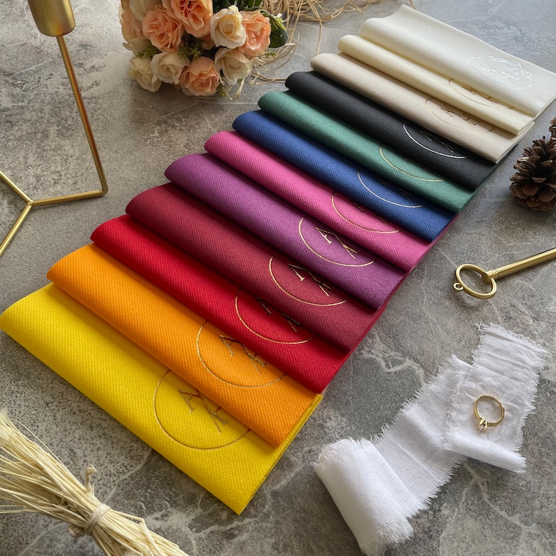 May include: A set of 12 folded napkins in various colours, including yellow, orange, red, maroon, purple, pink, blue, green, black, and white. Each napkin has a gold circle with a design in the centre.