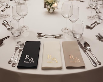 May include: A round white tablecloth set for a formal event. The table is set with glassware, silverware, and folded napkins in black, cream, and tan. Each napkin is embroidered with initials and a date.