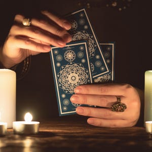 May include: Three rectangular tarot cards with a deep blue background and intricate white mandala-style geometric patterns. The cards are held upright, displaying detailed circular designs, surrounded by glowing candles in a dimly lit setting.