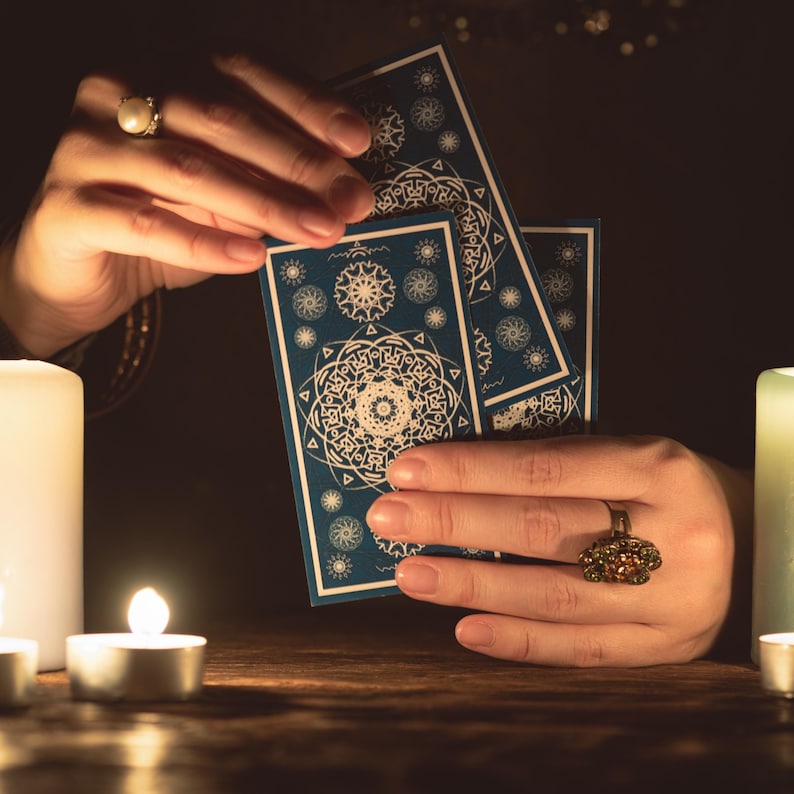 May include: Three rectangular tarot cards with a deep blue background and intricate white mandala-style geometric patterns. The cards are held upright, displaying detailed circular designs, surrounded by glowing candles in a dimly lit setting.
