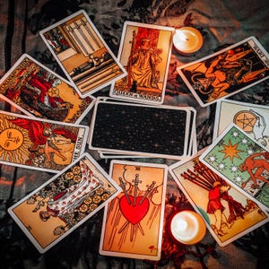 May include: A collection of tarot cards arranged in a circular pattern, including the Queen of Wands, The Devil, The Sun, and Three of Swords, with a central deck showing a dark starry back design and two small lit tea light candles.
