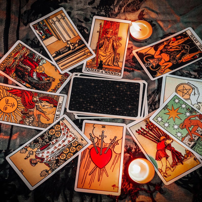 May include: A collection of tarot cards arranged in a circular pattern, including the Queen of Wands, The Devil, The Sun, and Three of Swords, with a central deck showing a dark starry back design and two small lit tea light candles.