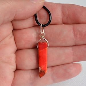 May include: A red and orange gemstone pendant on a black cord necklace. The pendant is a long, thin, irregular shape.