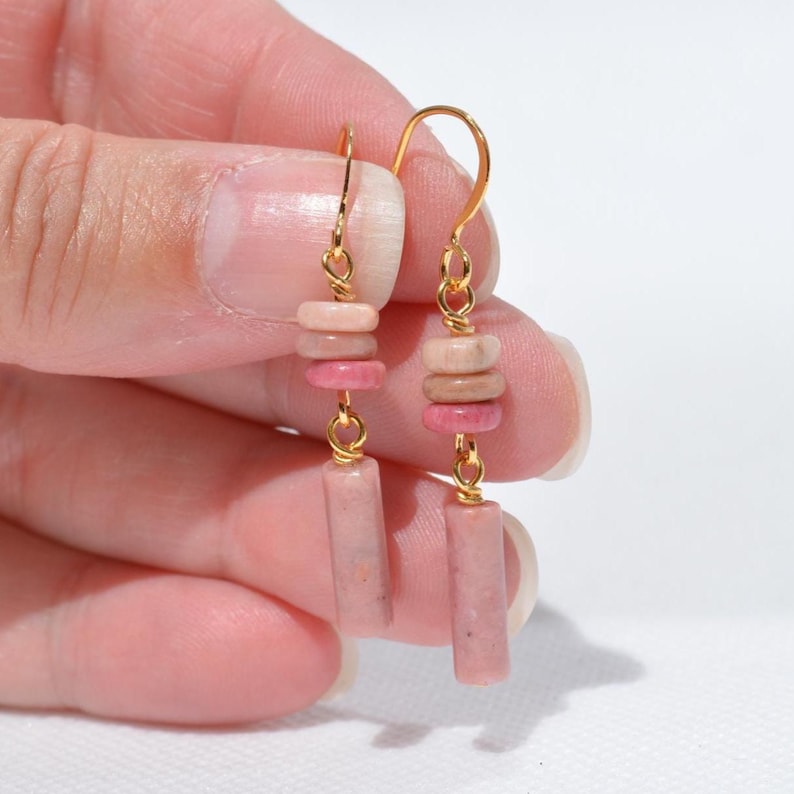 May include: A pair of gold-tone hook earrings featuring dangling pink stone beads. Each earring consists of three small, flat, circular pink stone discs stacked above a single, longer, cylindrical pink stone bead, all connected by gold-tone wire.