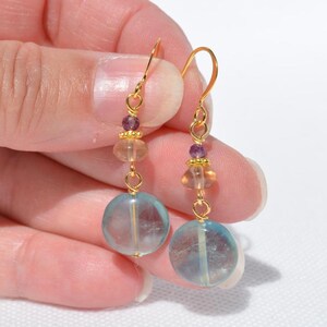 May include: A pair of gold-coloured dangle earrings. Each earring features a large, round, light blue bead at the bottom, with smaller orange and purple beads above. The earrings are held together with gold-coloured wire.