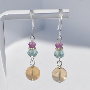 May include: A pair of dangle earrings with silver-tone hooks. Each earring features a light orange sphere, a light blue faceted bead, a cluster of pink beads, and silver-tone spacers. The earrings are displayed against a white background.