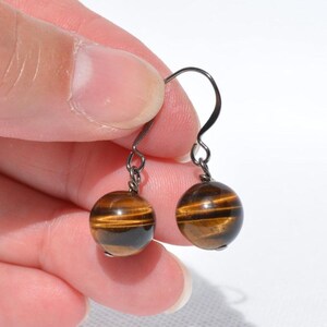 Puede incluir: Un par de pendientes colgantes con piedras preciosas de ojo de tigre esf&eacute;ricas con bandas de color marr&oacute;n dorado caracter&iacute;sticas. Las cuentas redondas y pulidas cuelgan de ganchos de metal oscuro, mostrando una superficie lisa y brillante.