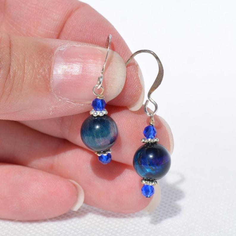 Op de afbeelding: Een paar hangende oorbellen met donkerblauwe, iriserende bolvormige kralen, geaccentueerd door kleinere, levendig blauwe gefacetteerde kristalkralen en zilverkleurige metalen tussenstukjes, hangend aan eenvoudige zilverkleurige oorhaken.