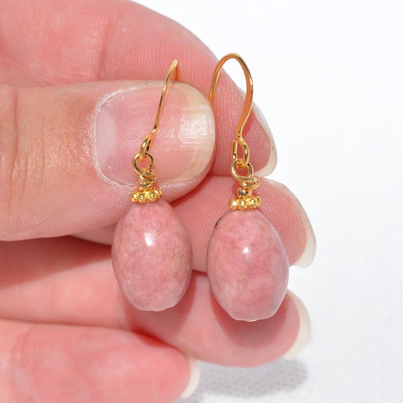 Peut inclure: Une paire de boucles d'oreilles pendantes compos&eacute;es de pierres de rhodonite rose lisses de forme ovale, suspendues &agrave; des crochets en m&eacute;tal dor&eacute;. Chaque pierre est surmont&eacute;e d'une petite perle dor&eacute;e textur&eacute;e au point de fixation.