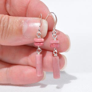 May include: A pair of drop earrings featuring silver-toned metal hooks and wire connectors. Each earring consists of two small, round, pink stone beads stacked above a single, longer, cylindrical pink stone bead, creating a vertical drop design.