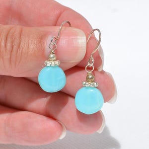 May include: A pair of drop earrings featuring round, opaque light blue beads suspended from silver-toned metal hooks. Each earring includes a small, decorative silver-toned metal bead cap with a scalloped edge positioned above the blue bead.