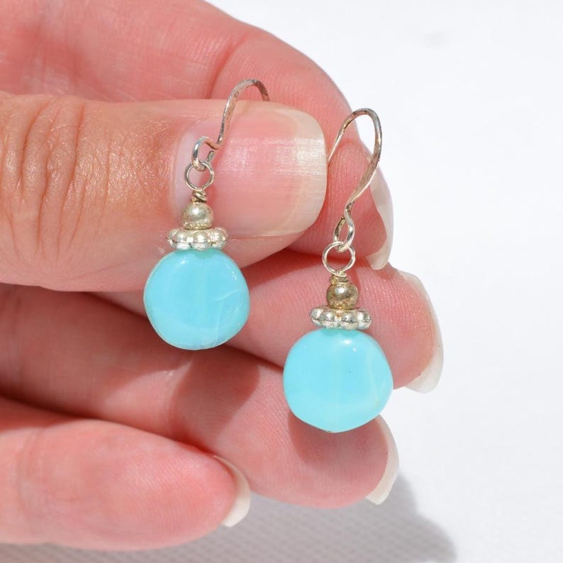May include: A pair of drop earrings featuring round, opaque light blue beads suspended from silver-toned metal hooks. Each earring includes a small, decorative silver-toned metal bead cap with a scalloped edge positioned above the blue bead.
