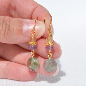 May include: Dangle earrings with gold-coloured hooks and a series of colourful beads. The earrings feature yellow, purple, and green beads. The bottom bead is a large, round, green and purple stone, suspended from the gold hook.