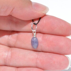 May include: A small, oval-shaped, light purple gemstone pendant. The gemstone is suspended from a silver-colored metal loop and wire, attached to a black cord. The pendant is held against a light background.