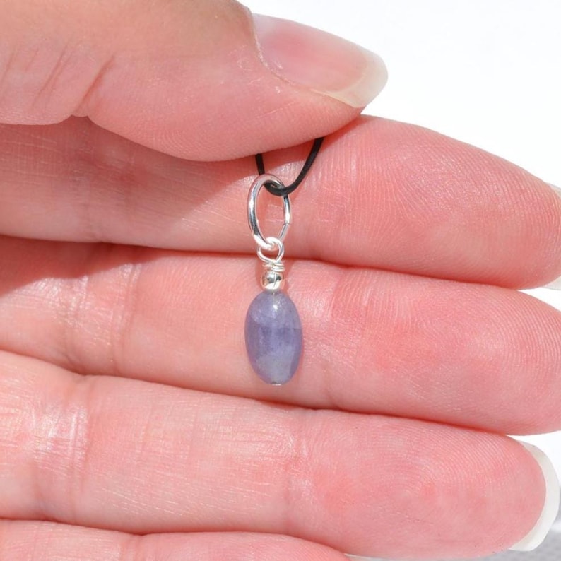 May include: A small, oval-shaped, light purple gemstone pendant. The gemstone is suspended from a silver-colored metal loop and wire, attached to a black cord. The pendant is held against a light background.