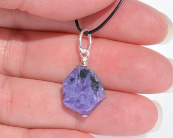 May include: A purple gemstone pendant with black inclusions, suspended from a black cord and silver bail. The raw, faceted stone is held in a hand, showcasing its natural texture and color variations. A small, silver bead accents the bottom.