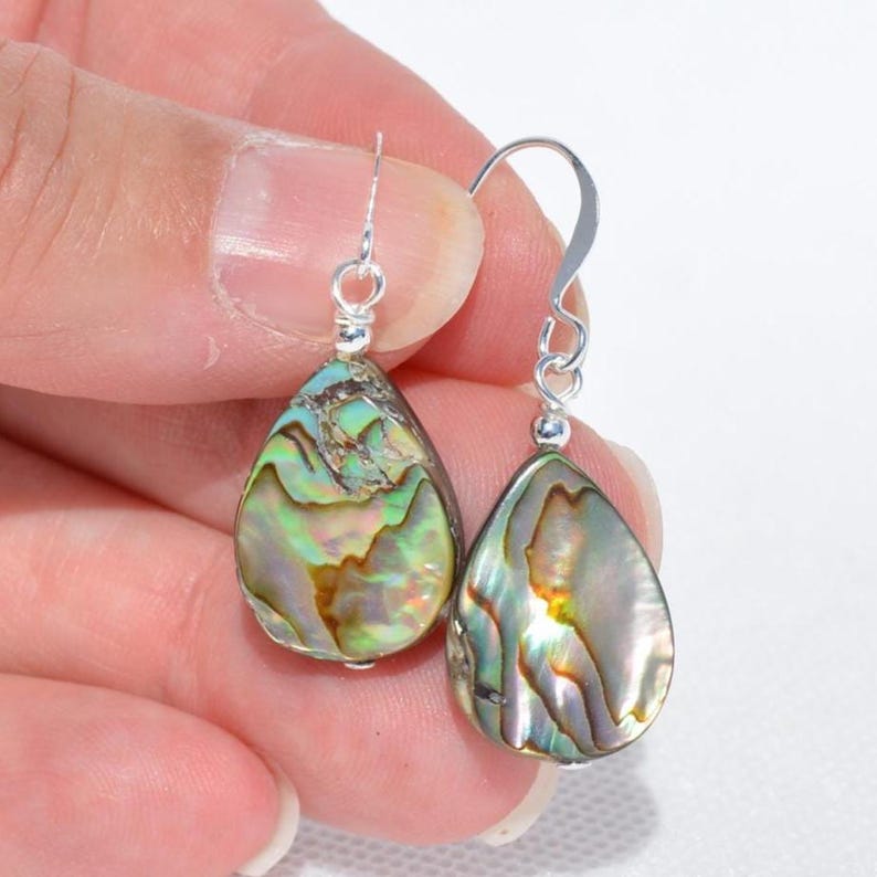 May include: A pair of teardrop-shaped earrings with iridescent abalone shells. The earrings have silver-colored hooks and small, decorative silver accents. The abalone shells display a mix of green, blue, and gold colors.