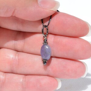 May include: A small, oval-shaped, lavender-colored gemstone pendant. The pendant is suspended from a black cord and features dark metal accents. The gemstone is approximately 0.75 inches in length, with a polished surface.