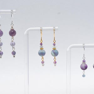 May include: Three pairs of dangle earrings with purple and blue beads. The earrings are displayed on white stands against a white background. The earrings have silver and gold-colored hooks and accents. The beads vary in size and shape, creating a layered effect.