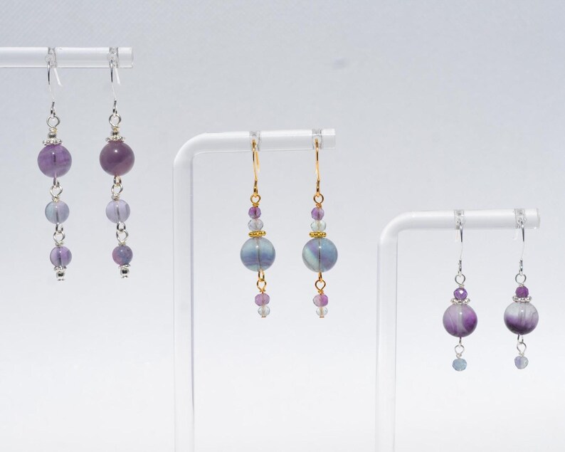 May include: Three pairs of dangle earrings with purple and blue beads. The earrings are displayed on white stands against a white background. The earrings have silver and gold-colored hooks and accents. The beads vary in size and shape, creating a layered effect.