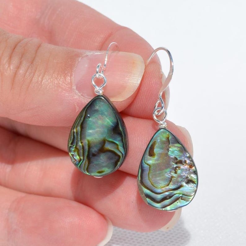 May include: A pair of teardrop-shaped earrings with iridescent abalone shells. The earrings have silver-colored hooks. The abalone shells display a mix of green, blue, and brown colors, with a pearlescent sheen.