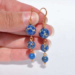 May include: Dangle earrings featuring three blue and tan jasper beads suspended from gold-colored hooks. The beads are round and vary slightly in size, with a marbled pattern. The earrings are held up by a hand.