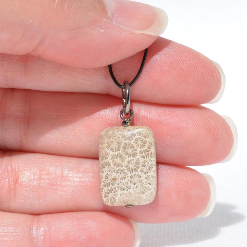 Op de afbeelding: Een rechthoekige hanger met een beige versteende koraalsteen met een gedetailleerd bloemachtig patroon. De steen hangt aan een donkere metalen ring die is bevestigd aan een dun zwart koord, met natuurlijke texturen en aardse tinten.