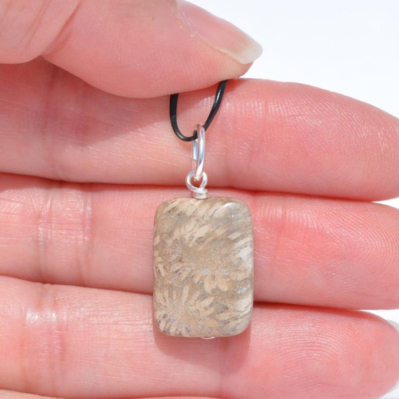 Op de afbeelding: Een rechthoekige hanger met een beige en bruin fossiel koraalpatroon met een gladde, gepolijste afwerking. De steen is bevestigd aan een zilverkleurig metalen oogje en hangt aan een dun, zwart koord, wat zorgt voor een eenvoudig, neutraal sieraad.