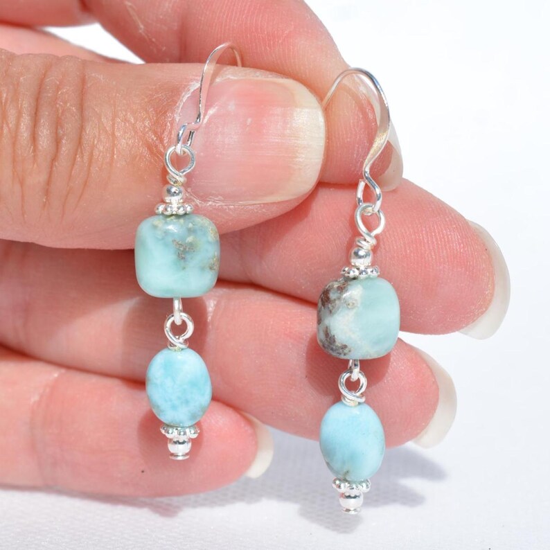 May include: A pair of dangle earrings featuring light blue and silver accents. Each earring has a silver hook, a square-shaped bead, a round bead, and decorative silver bead caps. The earrings are displayed against a white background.