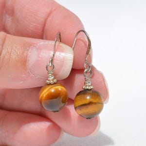 Puede incluir: Un par de pendientes colgantes con piedras preciosas de ojo de tigre redondas y pulidas, con bandas chatoyantes de color marr&oacute;n dorado. Cada piedra est&aacute; suspendida de un gancho de metal plateado mediante una peque&ntilde;a cuenta decorativa.