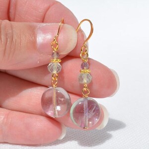 May include: A pair of gold-coloured dangle earrings. Each earring features a clear, round, iridescent bead at the bottom, with smaller clear and purple beads above. The earrings are held together with gold-coloured wire.