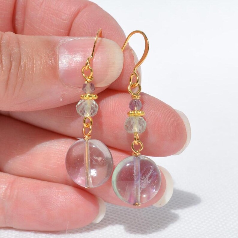 May include: A pair of gold-coloured dangle earrings. Each earring features a clear, round, iridescent bead at the bottom, with smaller clear and purple beads above. The earrings are held together with gold-coloured wire.