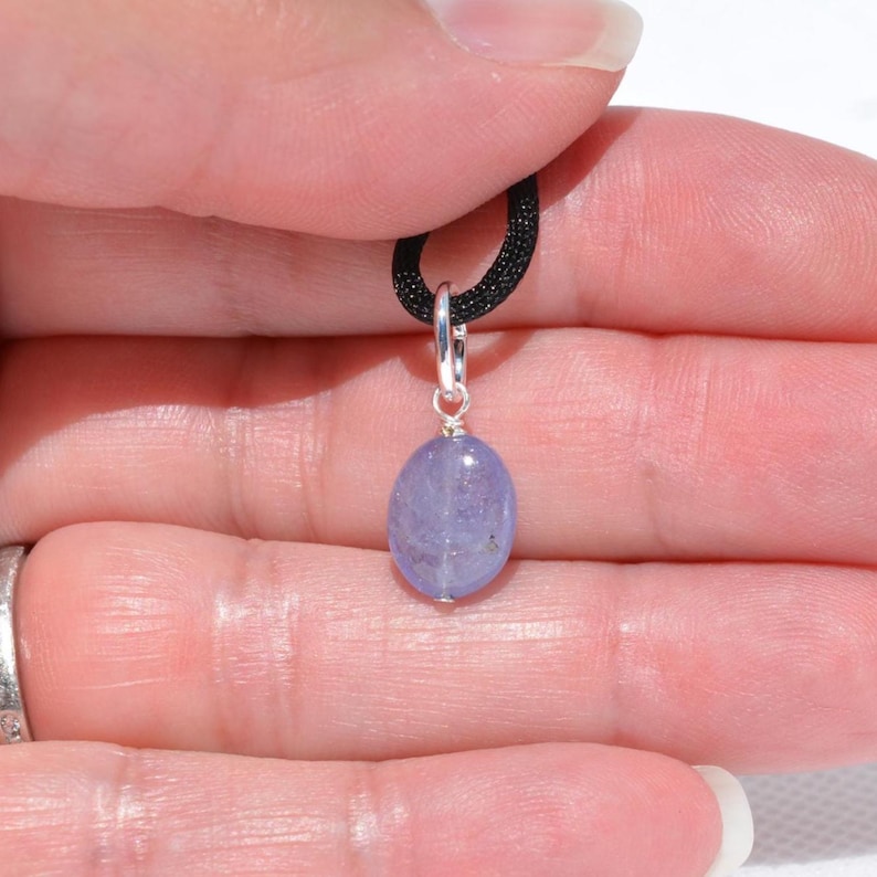 May include: A small, oval-shaped, light purple pendant with a silver bail hangs from a black cord. The pendant is a smooth, polished stone. The background is a person's hand.