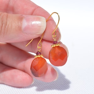 May include: Gold dangle earrings with a faceted carnelian stone and a small garnet bead at the top of the earring.