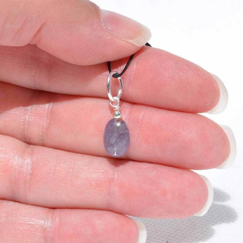 May include: A small, oval-shaped, lavender-colored pendant with a silver-colored bail and a black cord. The pendant is held by a hand against a white background. The pendant is a gemstone.