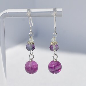May include: A pair of dangle earrings with a silver hook. Each earring features a large, round, vibrant purple bead at the bottom, connected by a silver wire to a faceted purple bead, a small light green bead, and a silver bead. Jewelry.