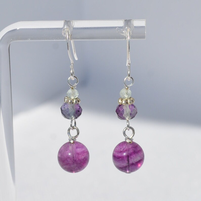 May include: A pair of dangle earrings with a silver hook. Each earring features a large, round, vibrant purple bead at the bottom, connected by a silver wire to a faceted purple bead, a small light green bead, and a silver bead. Jewelry.