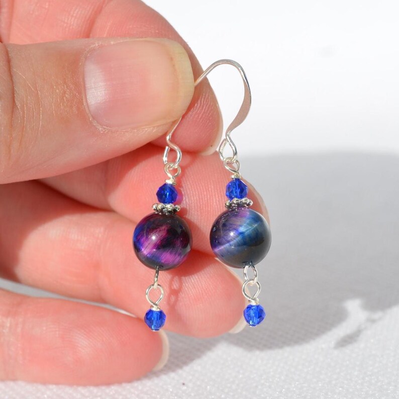 Op de afbeelding: Een paar hangende oorbellen met grote, ronde, iriserende tijgeroogkralen in paars en blauw. Elke oorbel is versierd met een kleine, gefacetteerde, felblauwe kristalkraal boven en onder de hoofdsteen, verbonden door zilverkleurige metalen onderdelen.