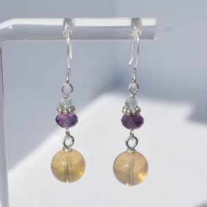May include: A pair of dangle earrings with silver hooks. Each earring features a large, round, yellow-orange bead at the bottom, a faceted purple bead, a small light blue bead, and silver accents. The earrings are displayed against a white background.