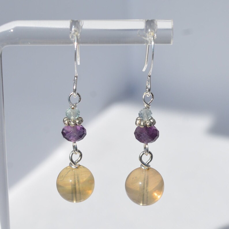 May include: A pair of dangle earrings with silver hooks. Each earring features a large, round, yellow-orange bead at the bottom, a faceted purple bead, a small light blue bead, and silver accents. The earrings are displayed against a white background.