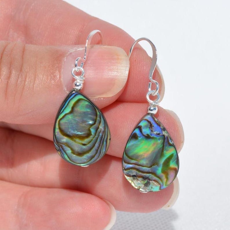 May include: A pair of teardrop-shaped earrings with iridescent abalone shells. The earrings have silver-colored hooks and small decorative beads. The abalone shells display a mix of green, blue, and gold colors.