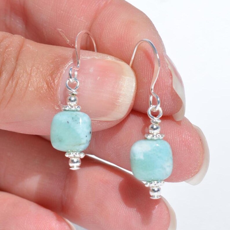 May include: A pair of dangle earrings featuring light blue-green, rectangular beads. The beads are suspended from silver-colored hooks and are accented with small silver beads at the top and bottom.