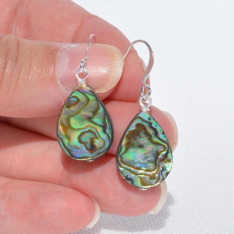 May include: A pair of teardrop-shaped earrings with iridescent abalone shells. The earrings have a silver-colored hook. The abalone shells display a mix of green, blue, and brown colors. The earrings are held by a person's hand.