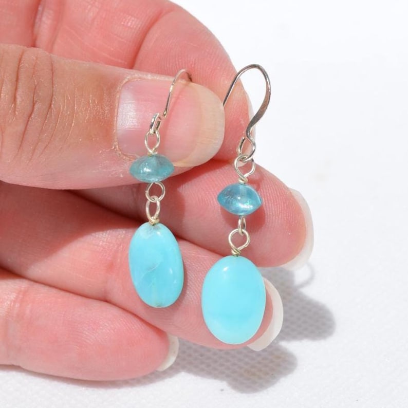 May include: A pair of drop earrings featuring silver-toned metal hooks, each suspending a small, translucent light blue bead above a larger, opaque, oval-shaped turquoise-coloured stone. The earrings are designed with simple wire links.