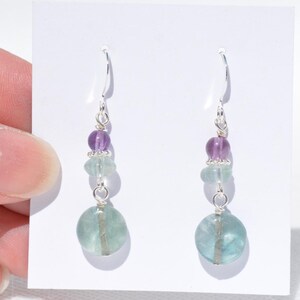 May include: Dangle earrings with purple and green beads. The earrings have silver hooks and wire, with a large, round, light green bead at the base. The beads are strung together, creating a layered effect. The earrings are designed to hang.